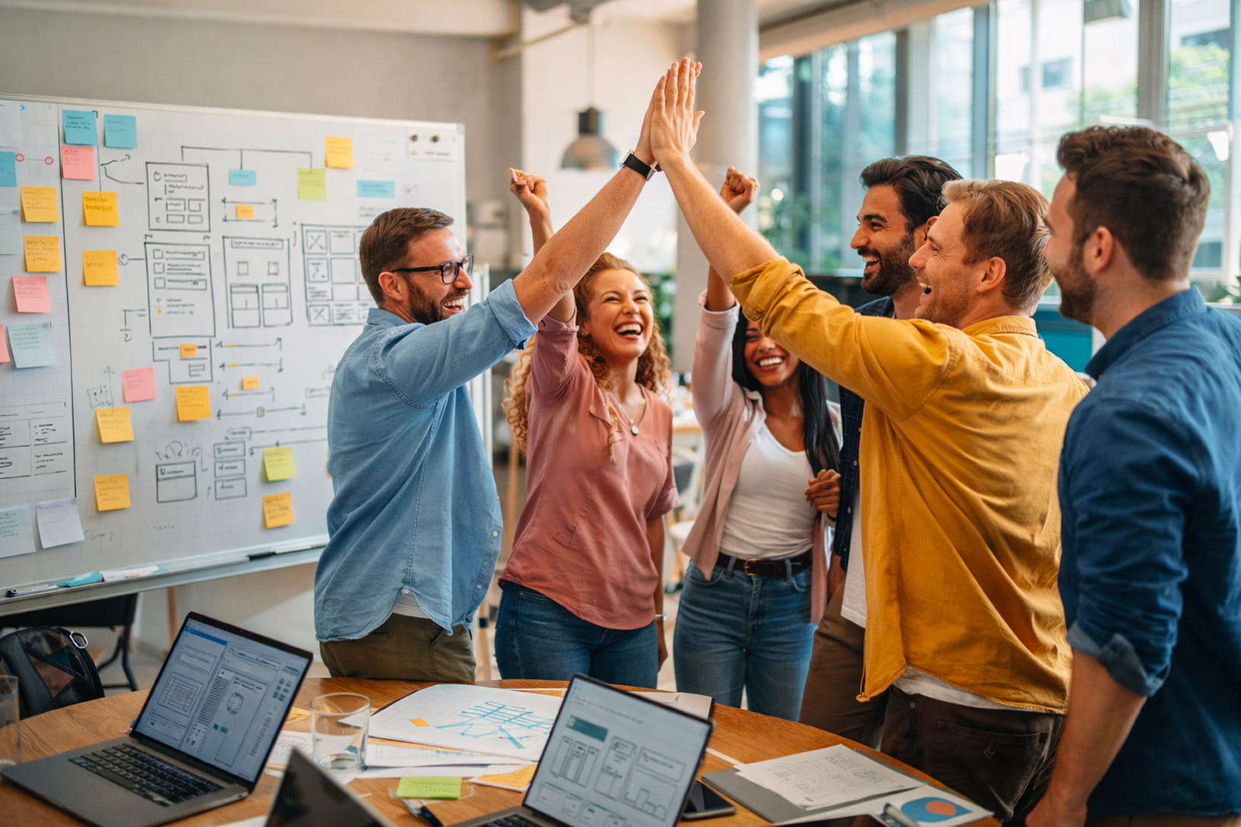 Digital agency team celebrating a new project kickoff around a workspace with laptops, website wireframes, and planning sketches on a whiteboard.